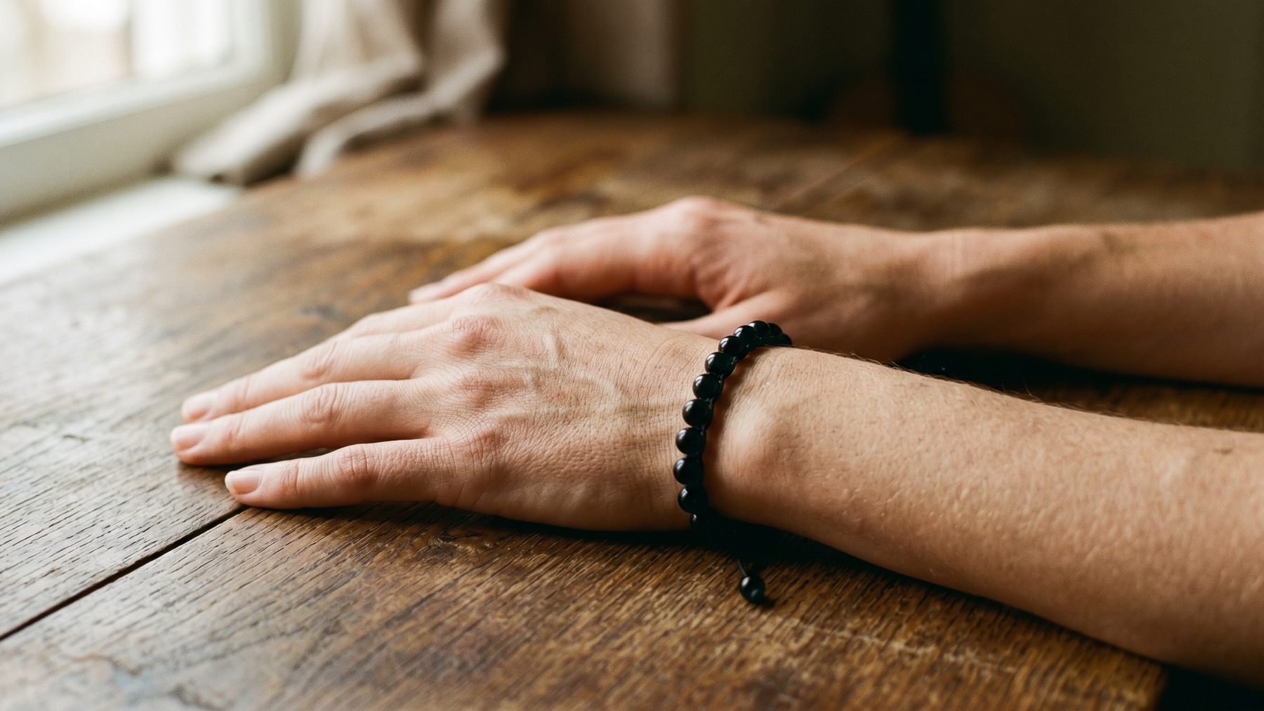 Open hands resting on a wooden table, obsidian bracelet visible, warm light